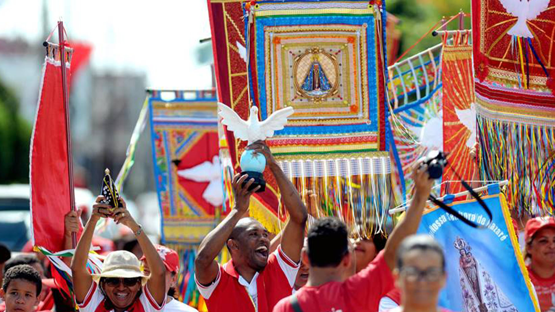 Imagem ilustrativa: fotografia de pessoas com roupas vermelhas de brancas andando na rua e segurando bandeiras com imagens de pombas brancas e Santa Maria.